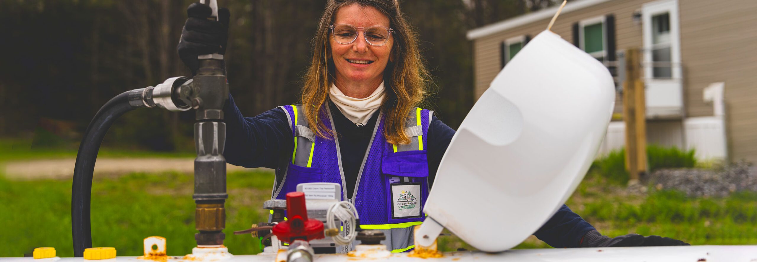 A female worker stands against an outdoor propane tank with a fueling hose attached.