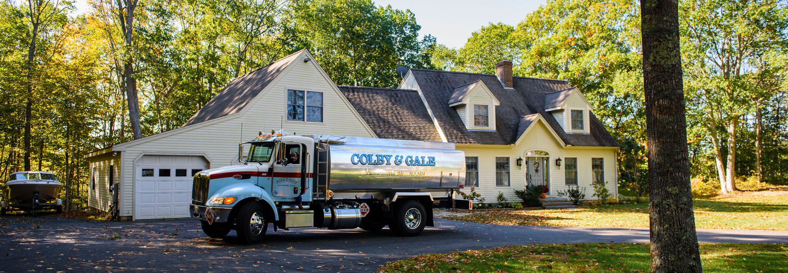 A shiny heating oil truck parked in front of a yellow 'Cape Cod' style home surrounded by trees.