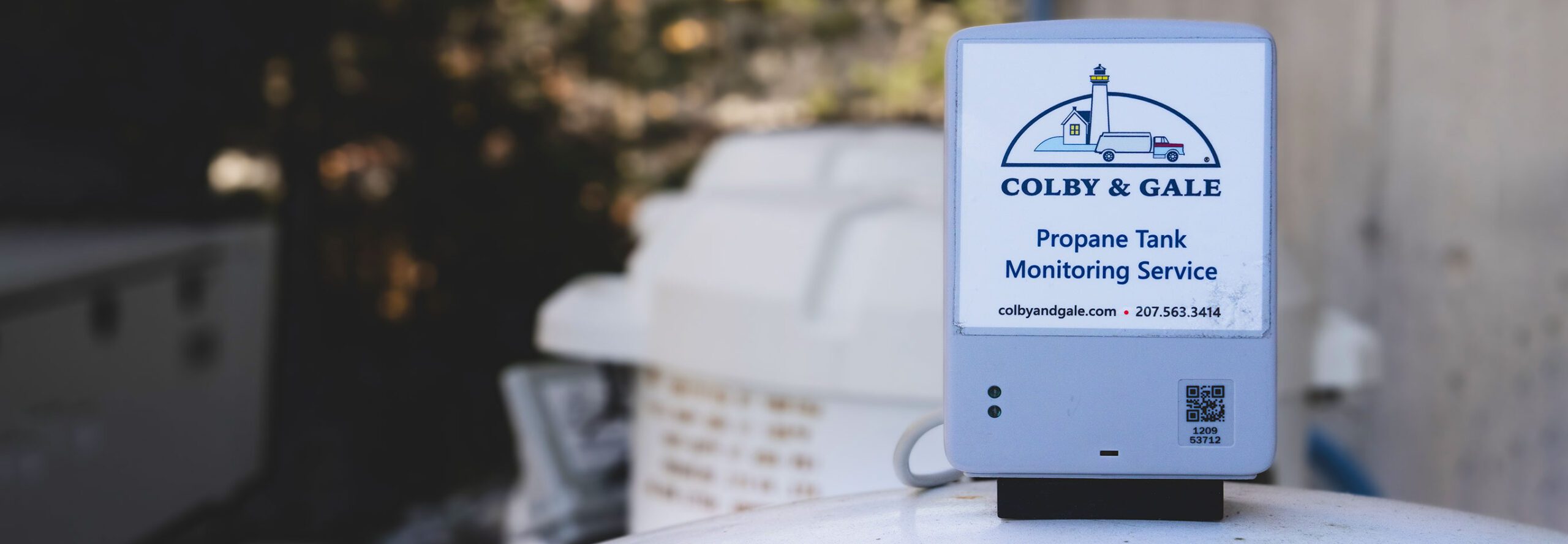 A gray box sits on top of a white propane tank outside of a Maine home.