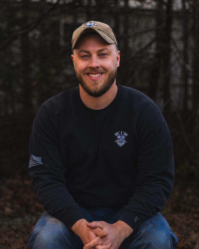Bryce Collamore sitting wearing a hat with hands crossed and a military sweatshirt smiling.