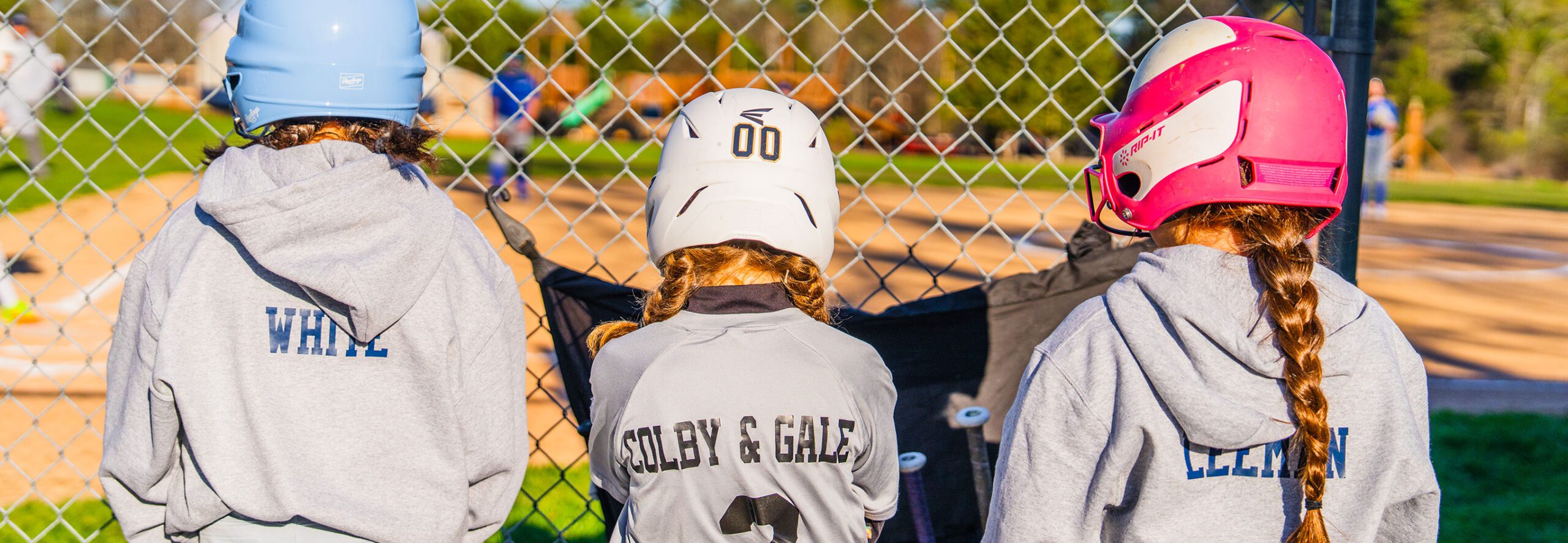 Three girls wearing softball helmets stand in a row watching the game