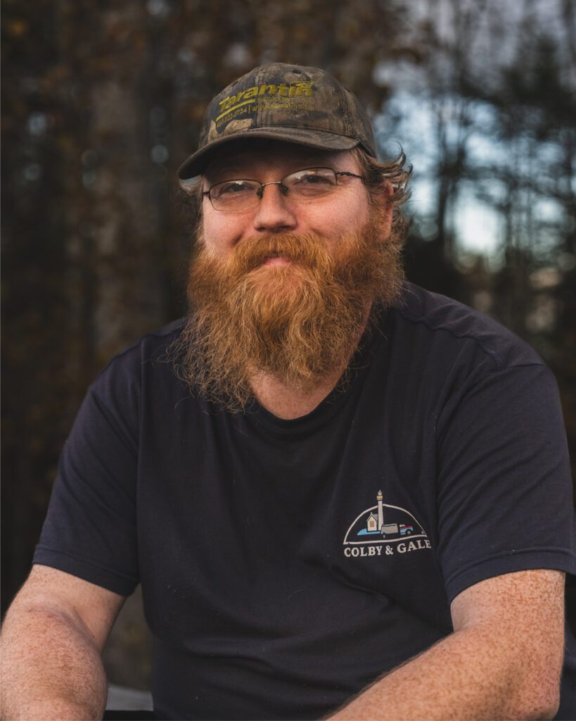 Cory Lincoln sitting and wearing a hat with beard and colby & gale shirt. 