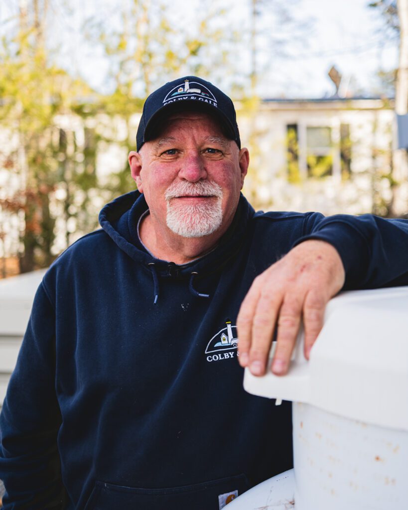 Darly Hodgkins wearing Colby & Gale hat and sweatshirt with left arm resting on top of propane tank.