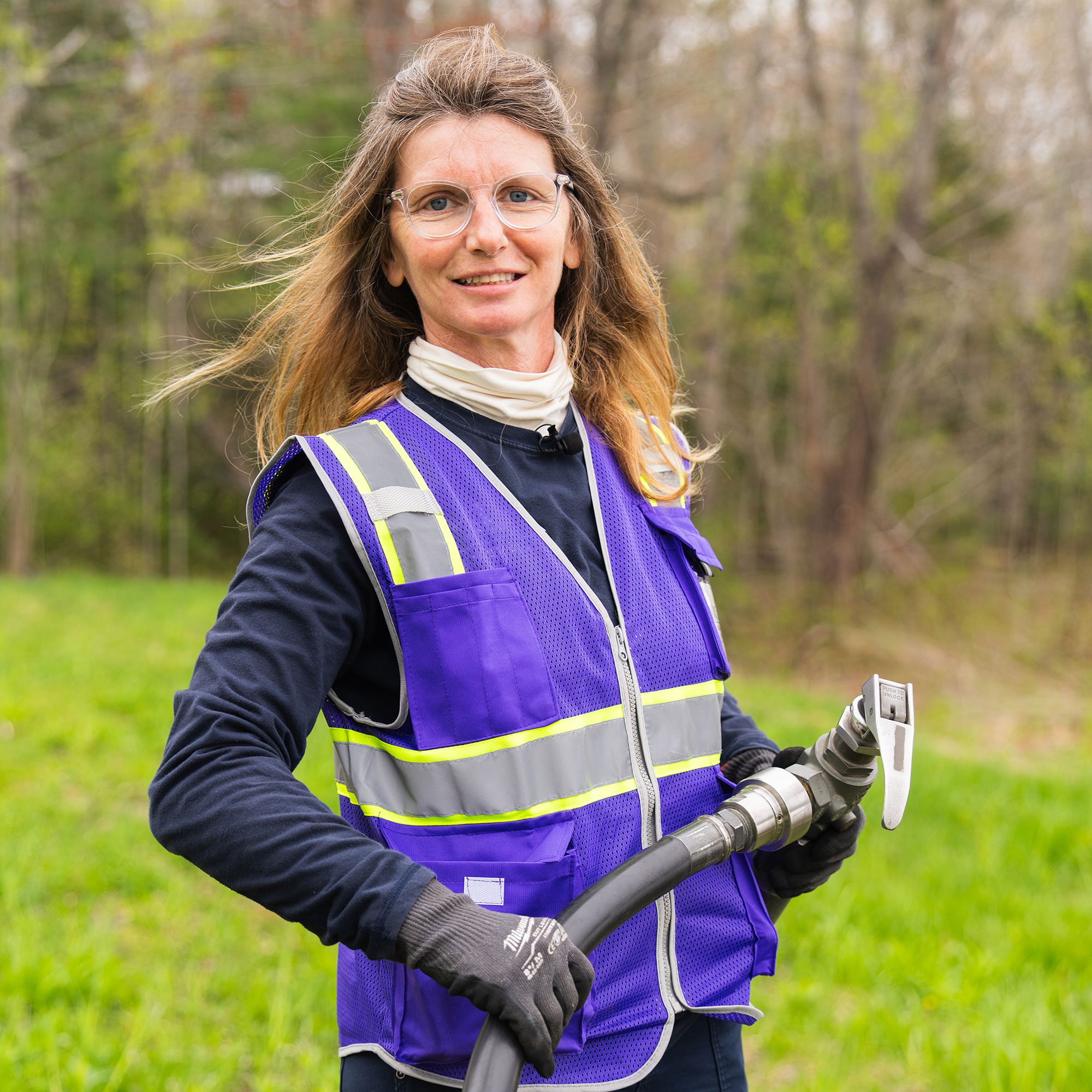 A woman in a purple reflector vest and glasses holds a fuel hose.