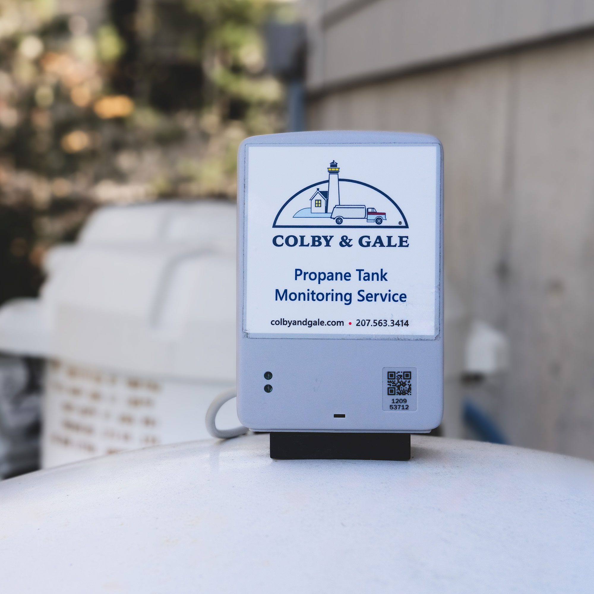 A small gray box with the words propane tank monitoring service sits on top of a propane tank.