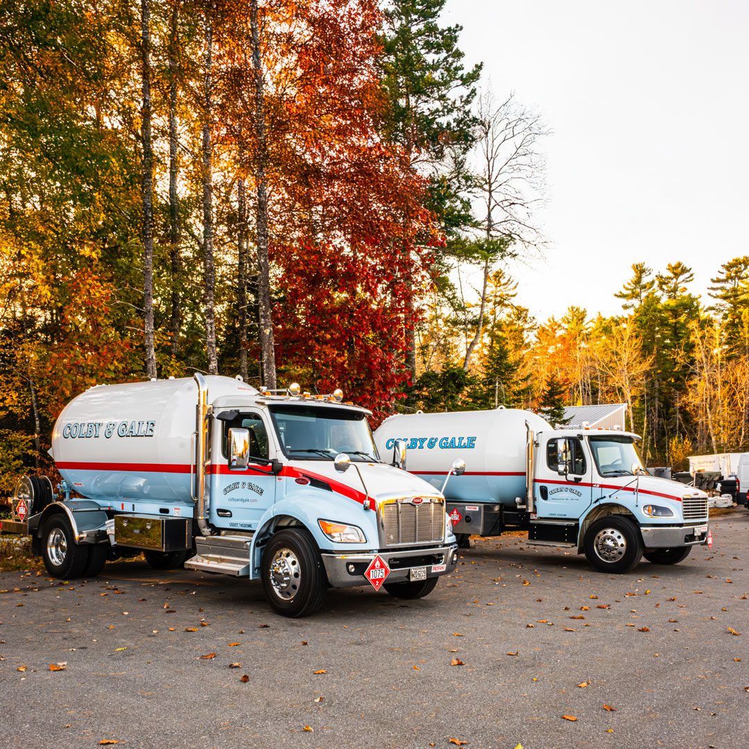 Two white and blue pill-shaped propane trucks parked against colorful new england foliage.